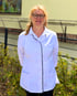 Lächelnde Frau mit Brille und blonden Haaren im weißen Kittel vor einem gelben Gebäude und grünen Pflanzen.