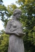 Verwitterte Steinstatue einer Frau mit geflochtenen Haaren, die eine Schale in den Händen hält, vor unscharfen grünen Bäumen.