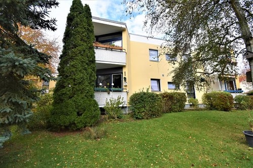 Mehrfamilienhaus mit gelber Fassade, Balkonen und bewachsenem Garten mit Bäumen und Sträuchern.