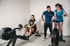 Zwei Männer und eine Frau trainieren in einem Fitnessstudio an Rudergerät und Air Bike, ein Trainer assistiert.