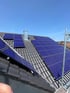 Dunkle Dachziegel mit blauen Solarmodulen und einem Schornstein unter blauem Himmel.