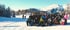 Eine große Gruppe von Skifahrern posiert auf einer verschneiten Piste vor einem Bergpanorama bei strahlendem Sonnenschein.