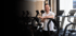 Lächelnder Mann in weißem Polohemd lehnt sich an ein Fitnessgerät in einem Fitnessstudio mit weiteren Geräten.