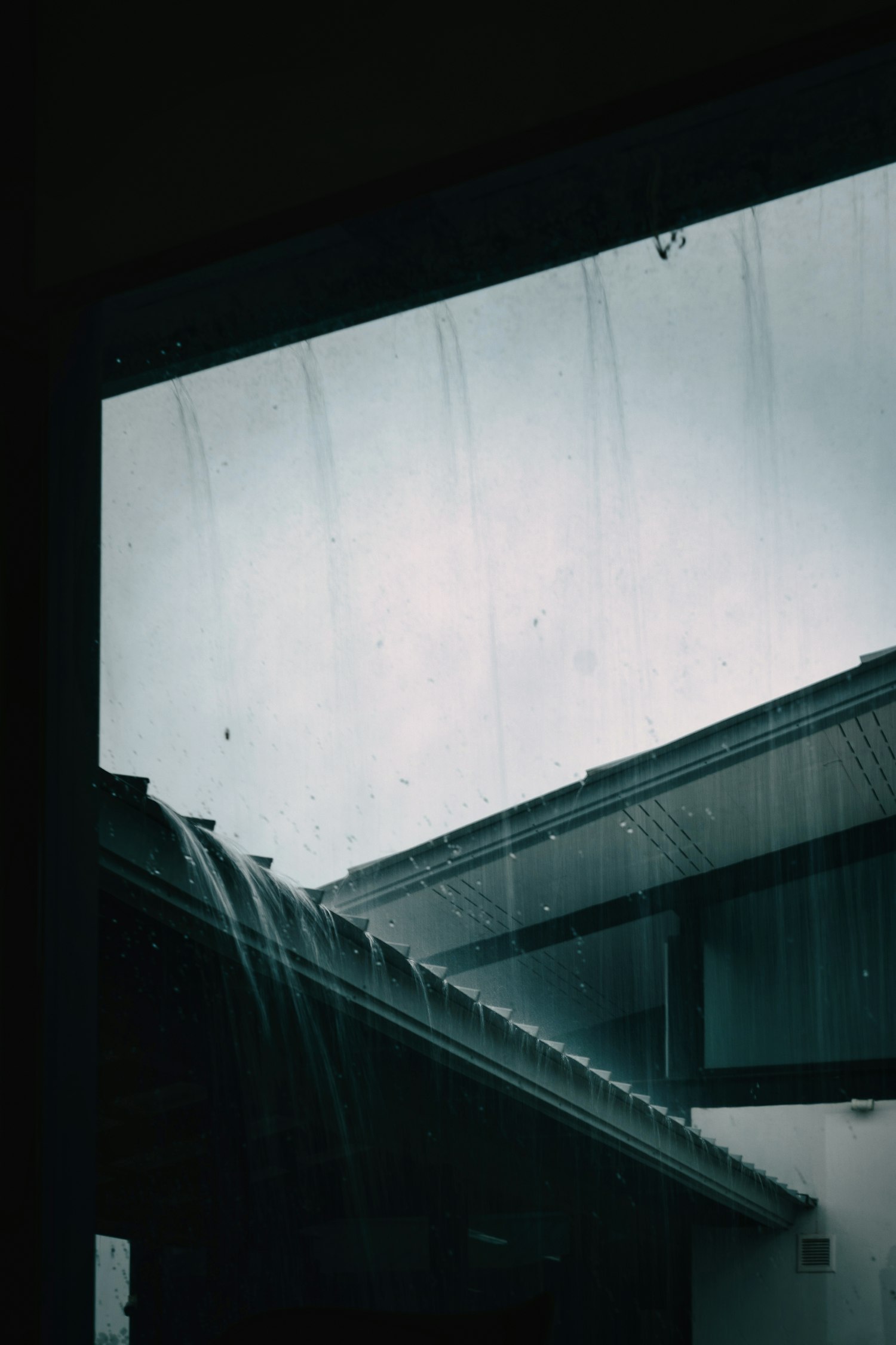Heavy rain running off a roof edge past a window