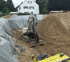 Bagger in einer Baugrube mit Erdhaufen und grauer Plane.