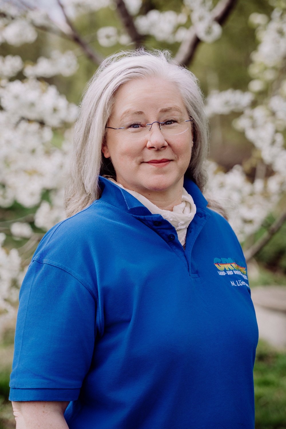 Frau mit grauem Haar und Brille trägt ein blaues Poloshirt mit buntem Logo und steht vor blühenden weißen Blumen.