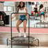 Eine lächelnde Frau springt auf einem Trampolin in einem Fitnessstudio, zwei weitere Frauen springen im Hintergrund.