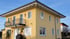 Gelbes zweistöckiges Einfamilienhaus mit Walmdach, weißem Dachüberstand und Balkon mit schwarzem Schmiedeeisengeländer.