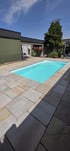 Rechteckiger Außenpool mit hellblauem Wasser, umgeben von einer Terrasse aus beigen Natursteinplatten im Garten.