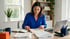 A woman in a blue shirt reviews paperwork at a desk with a laptop, notebook, and office supplies in a bright room.
