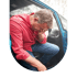 A man in a red shirt sits in a blue car with his head in his hand, appearing distressed or fatigued.