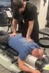 A chiropractor in black scrubs performs a manual spinal adjustment on a patient lying face down on a chiropractic table.