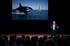 Man in a suit speaks on stage to an audience, with a screen behind him showing an orca and a sailboat.