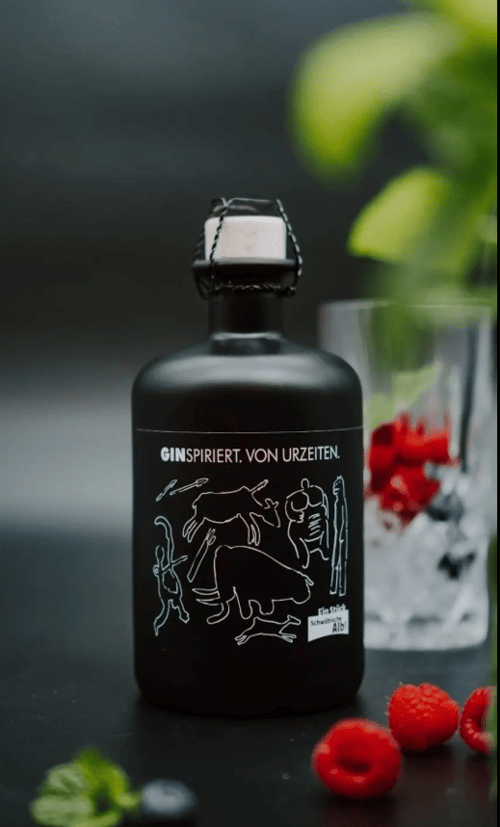 Flasche des Hausgins ‚Ginspiriert. Von Urzeiten.‘ mit Höhlenmalerei-Motiv, daneben ein Glas mit Eis und Beeren