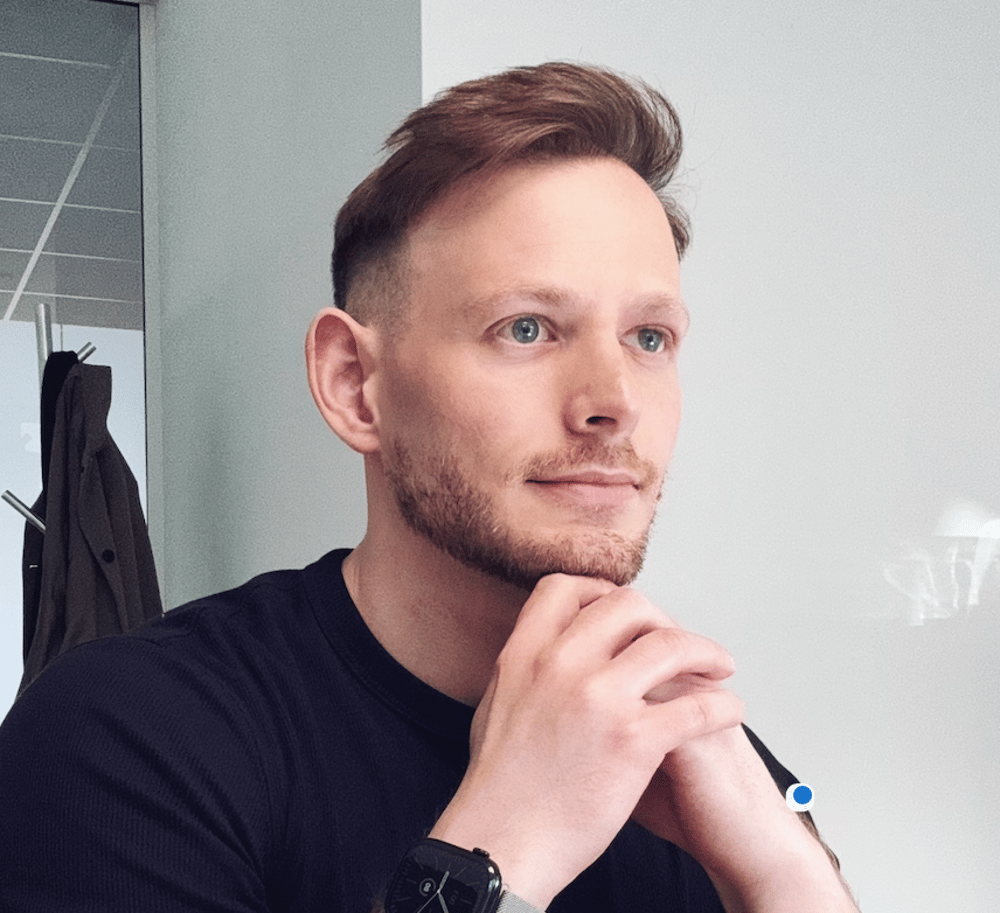 Ein lächelnder Mann mit Bart und Brille in einem grauen Sakko vor einem gelben Hintergrund.