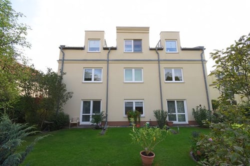 Beiges dreistöckiges Haus mit fünf Fenstern und zwei Glastüren im Erdgeschoss, umgeben von einem grünen Garten.