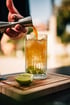 Eine Hand gießt orangefarbene Flüssigkeit aus einem Jigger in ein Glas mit Eis und grüner Flüssigkeit auf einem Holzbrett mit Limette.