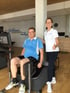 Ein Mann trainiert an einem Fitnessgerät, während eine lächelnde Trainerin daneben steht und den Daumen nach oben zeigt.