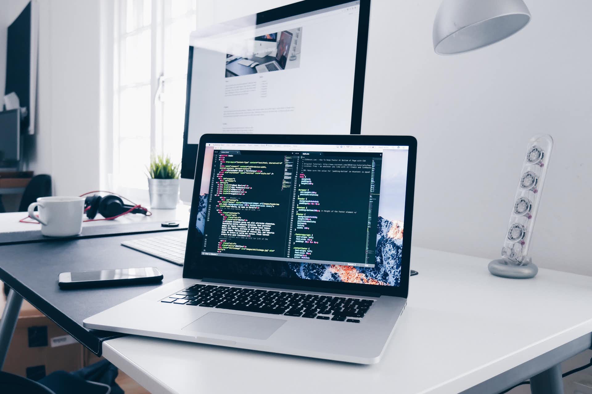 Laptop auf Schreibtisch mit Code auf dem Bildschirm, daneben ein größerer Monitor und eine Tasse Kaffee.