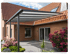 Moderne Terrassenüberdachung aus grauem Aluminium mit Glasdach an einem Backsteinhaus mit roten Ziegeln und Blumen.