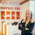 Eine Frau mit Headset und schwarzem Blazer steht lächelnd vor dem Empfangstresen der Praxis Physio CKI.