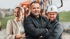 Drei lächelnde Arbeitskräfte stehen vor einer Baustelle mit orangefarbenem Kran, ein Mann trägt einen weißen Schutzhelm.
