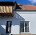 Weiße Hausfassade mit Holzbalkon, grauer Tür und Fenster mit Rollladen unter rotem Ziegeldach.