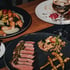 Drei Gourmetgerichte mit Fleisch, Lachs, Pasta, Gemüse, Couscous und einem Glas Weißwein auf einem Holztisch.