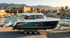White and navy motorboat on a trailer, parked on a stone dock with a harbor and historic city in the background.