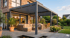 Moderne Pergola mit Lamellendach über einer Terrasse mit Lounge-Möbeln vor einem Haus mit großen Glasfenstern.