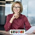 Lächelnde Frau mit Brille und roter Bluse am Schreibtisch; Logo der Verwaltungsgemeinschaft Memmingerberg im Vordergrund.