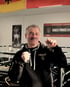 Älterer Mann im Hoodie mit geballten Fäusten im Boxring. Im Hintergrund eine Flagge und ein Poster von Muhammad Ali.