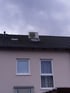 Eine Klimaanlage steht auf einem schwarzen Ziegeldach über zwei Fenstern einer hellrosafarbenen Hauswand.