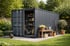 Dunkelgrauer Schiffscontainer als Gartenschuppen mit Regalen, Rasenmäher, Schubkarre und Gartengeräten im Grünen.