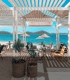 Ein Strandcafé mit dem Namen Beach Bar Buquez mit weißen Holzpergolen, Tischen, Stühlen, Pflanzen und Blick auf blaues Wasser, Strand und Inseln.