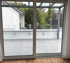 Glasschiebetüren öffnen sich zu einer modernen Terrasse mit Fliesenboden, Geländer und Blick auf Bäume und Häuser.