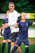 Ein lächelnder Trainer mit drei Fußballspielern in blauen Trikots auf einem grünen Feld.