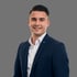 Nico Riedl, freundliches Business-Porträt eines jungen Mannes im blauen Anzug mit offenem weißen Hemd vor einem neutralen Hintergrund.