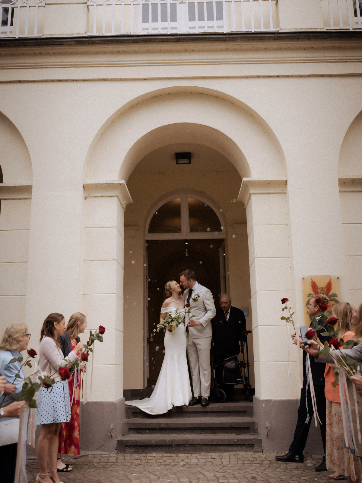 Brautpaar küsst sich im Eingangsportal, Gäste mit Rosen, am Schloss Eller in Düsseldorf.