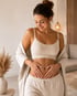 Lächelnde Frau in beiger Loungewear hält ihre Hände sanft auf ihren flachen Bauch in einem hellen Wohnzimmer.