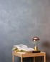 Graue Wand mit Putzstruktur, darauf ein Holztisch mit aufgeschlagenem Buch und einer modernen Lampe.