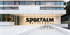 Modernes weißes Gebäude mit dem Schriftzug "SPORTALM KITZBÜHEL" über dem Eingang und einer reflektierenden Fläche davor.