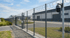 Ein hoher Sicherheitszaun mit Stacheldraht und Überwachungskamera vor einem modernen Industriegebäude mit Lagerhallen.