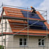 Ein Arbeiter installiert Solarmodule auf einem roten Ziegeldach eines Hauses mit Gerüst.