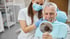 Eine Dentalhygienikerin mit Maske und Handschuhen zeigt einem lächelnden älteren Mann seine Zähne in einem Spiegel.