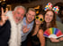 Vier lachende Partygäste: Ein Mann mit weißer Boa, ein Mann winkend, eine Frau mit Sternenbrille, eine Frau mit Regenbogenfächer.