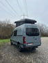 Heckansicht eines Mercedes Sprinter Reisewagens mit Aufstelldach – kompakter Wohnmobil-Ausbau mit grauer Lackierung und individuellem Heckausbau, fotografiert auf Kopfsteinpflaster vor bewaldeter Kulisse