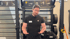 Ein Fitnesstrainer steht in einem Fitnessstudio vor Regalen mit Medizinbällen, Kettlebells und Gymnastikbällen.