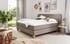 Graues Boxspringbett mit weißer Matratze, Kissen, Steppdecke und Nachttisch in einem Schlafzimmer.