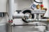 Schweißer mit Schweißhelm und Handschuhen schweißt Metallstück auf großem Gittertisch in einer Fabrikhalle.